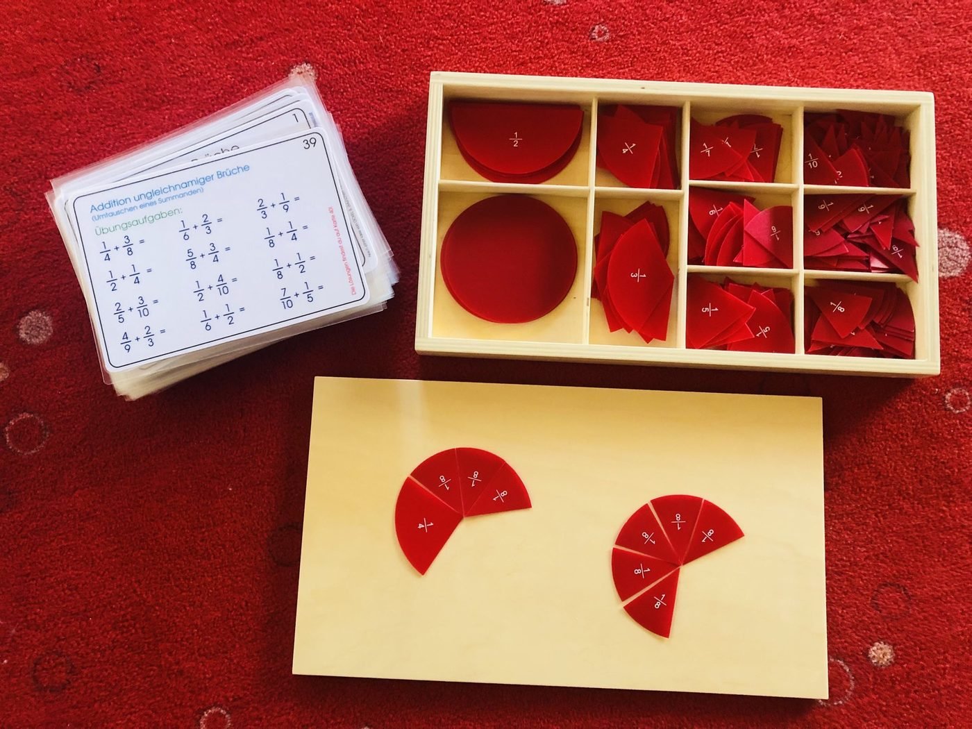 Montessori-Bruchrechenmaterial: rote Kreissegmente in einer Holzschachtel, daneben Aufgabenkarten zur Addition ungleichnamiger Brüche.
